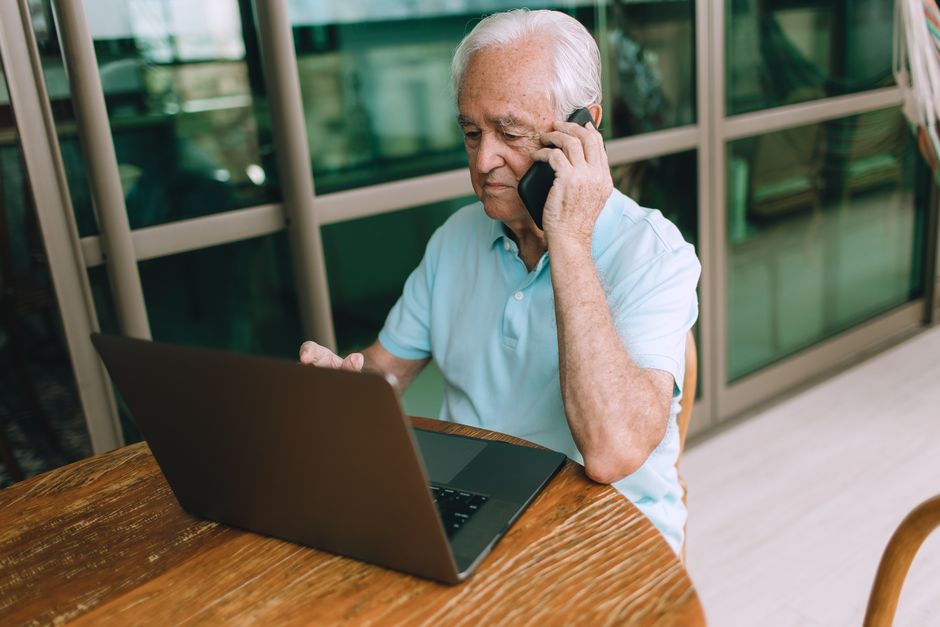 Elderly man multitasking, using laptop and phone at home. Lifestyle image.
