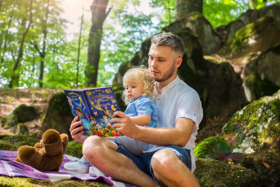 Father and toddler bonding over a book in a sunny forest setting.