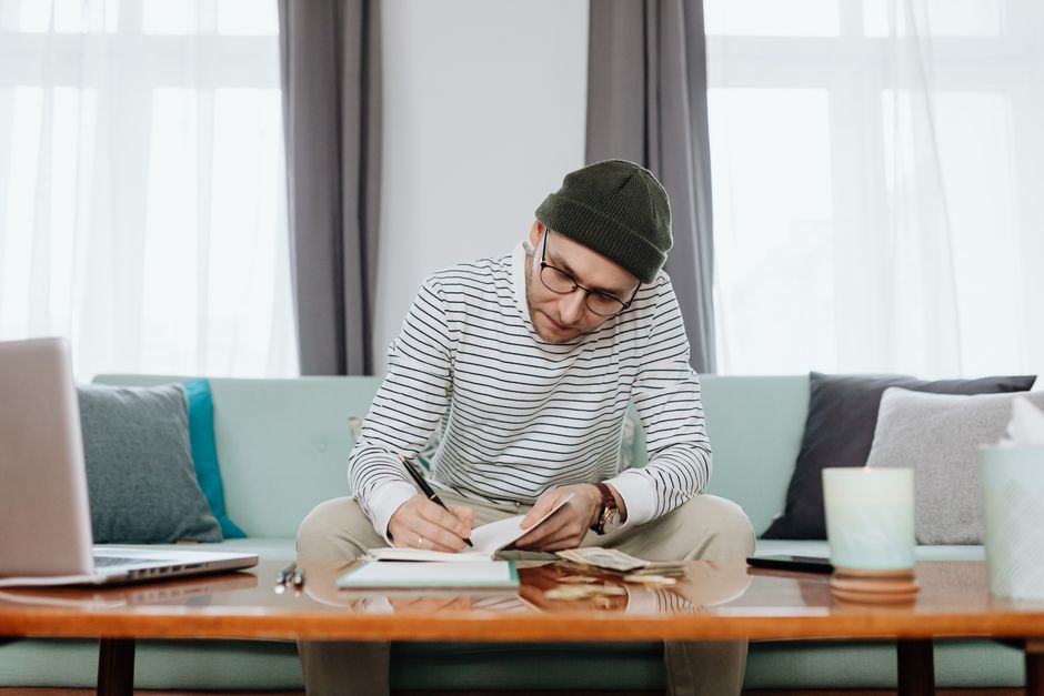 A man in a beanie writing notes and budgeting at a living room desk with a laptop.