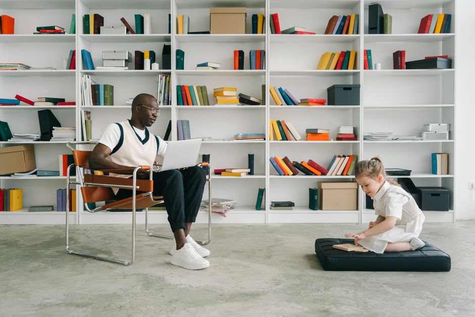 Man working on laptop while child reads in a modern library setting.