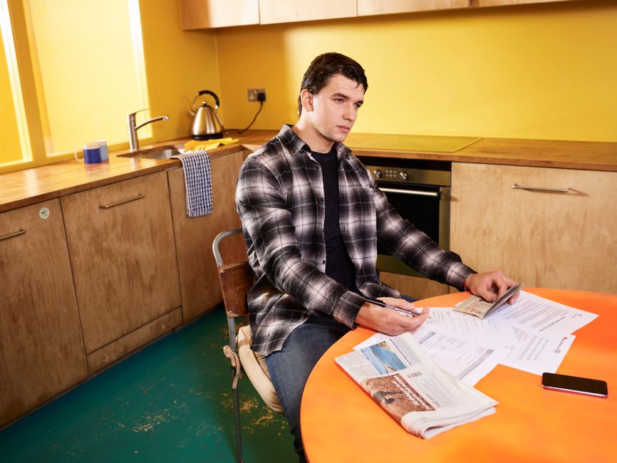 Man sitting in kitchen reviewing bills and a check with smartphone nearby.