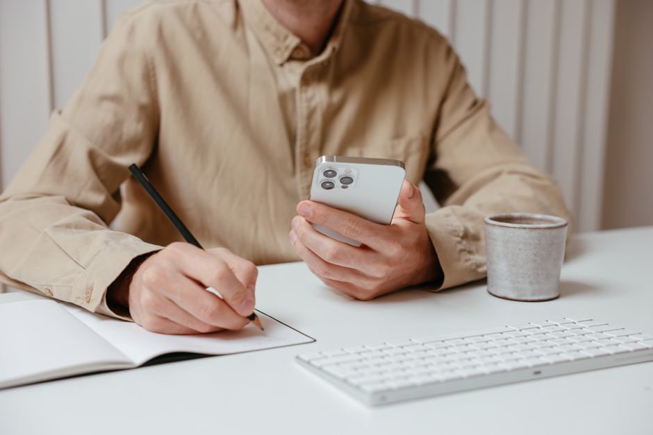 Person writing notes while holding a smartphone at a desk, showcasing multitasking.