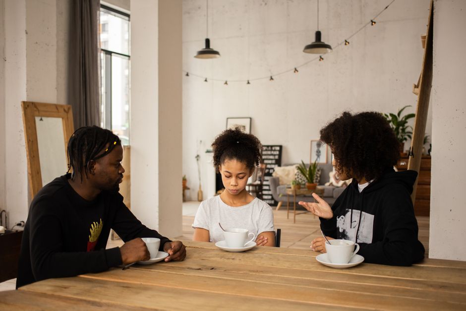 African American mother scolding unhappy daughter sitting at table with cups of coffee near father