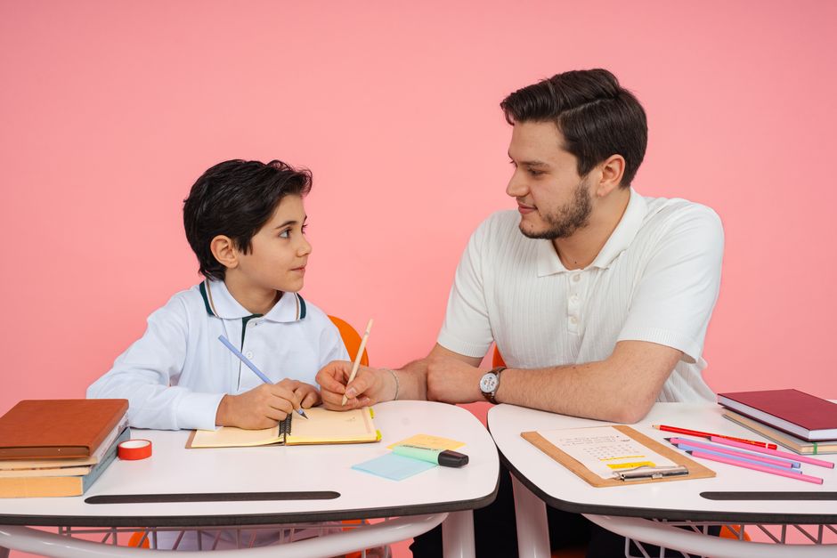 Father teaching his son at a desk with colorful stationery on a pink background.