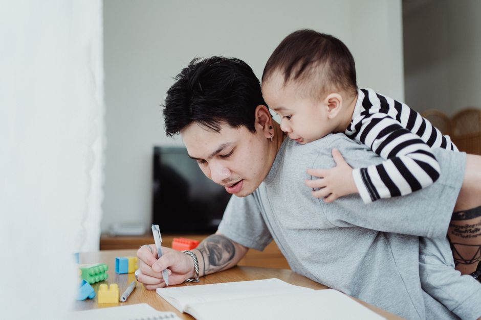 A father writing while his young son playfully clings to him indoors, showcasing fatherhood and bonding.