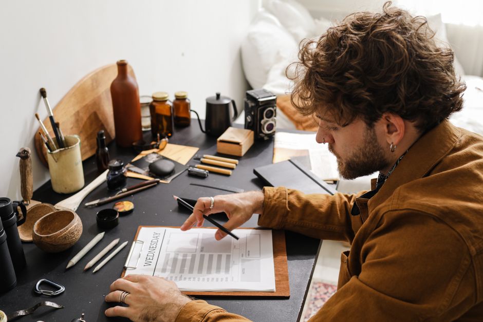 Man with curly hair planning at his desk, surrounded by notebooks and tools, in a cozy home office.
