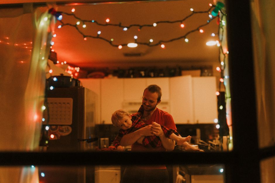 Father and son share a playful moment in a warmly lit kitchen decorated with holiday lights.